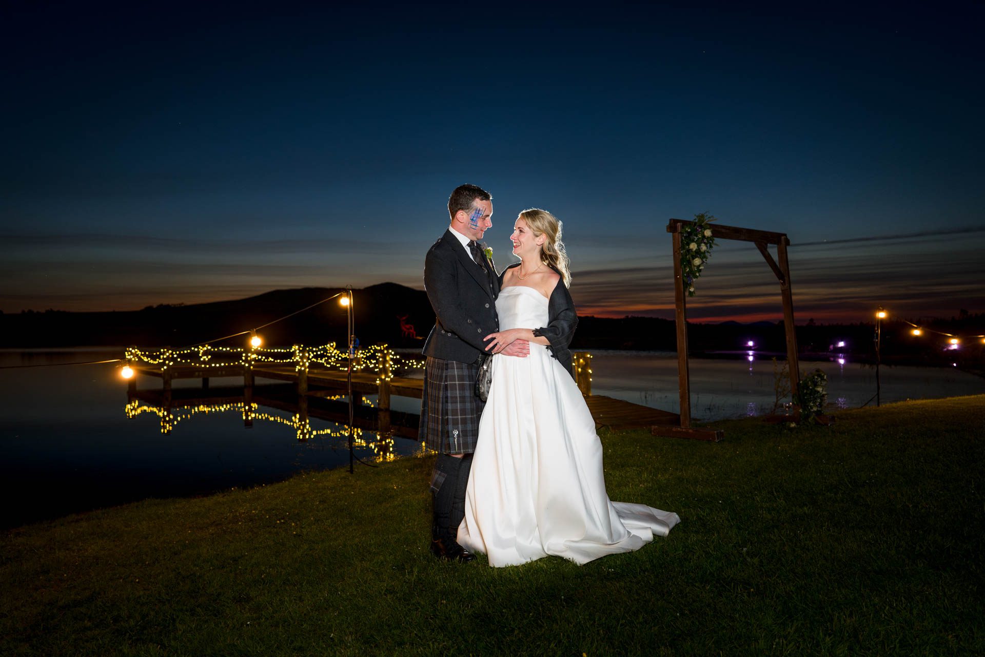 The closing portrait of Hollie and Colin at their Scottish Borders Wedding. Background of lake with jetty illuminated by fairy lights and the last glow of sunset in the sky. 