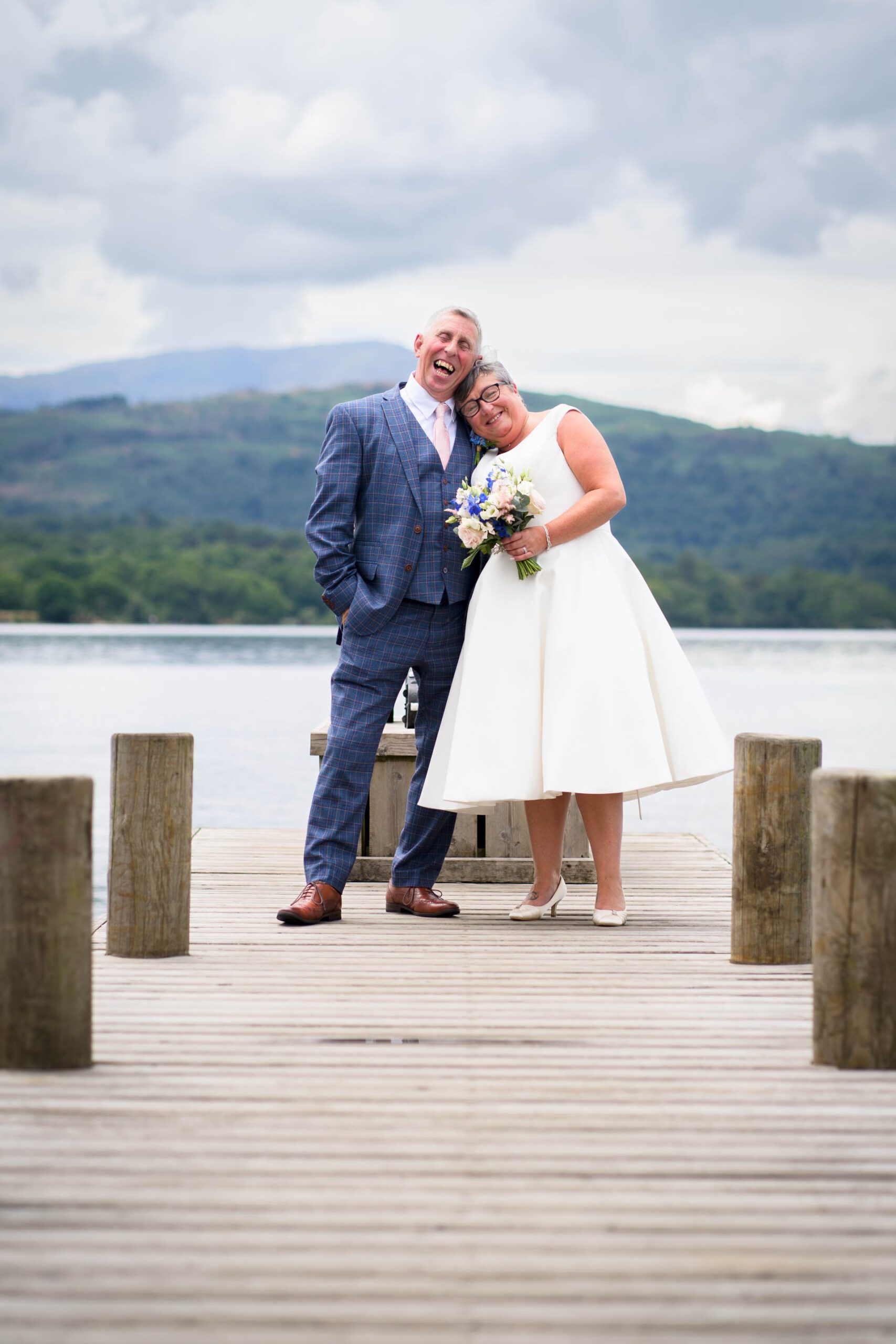 Wedding portrait of bride and groom on jetty at Windermere with lake and mountains in background. Taken at their Low Wood Bay Resort wedding.