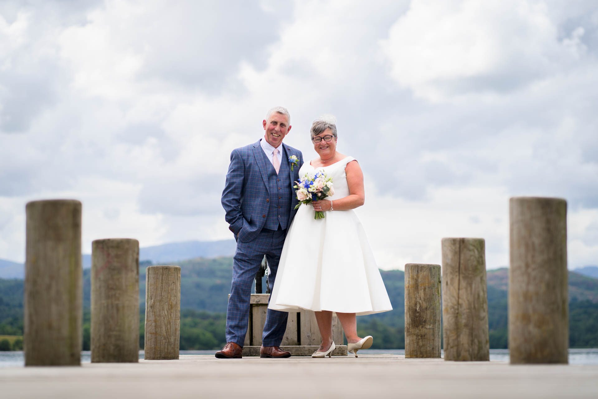 Wedding portrait of bride and groom on jetty at Windermere with lake and mountains in background. 