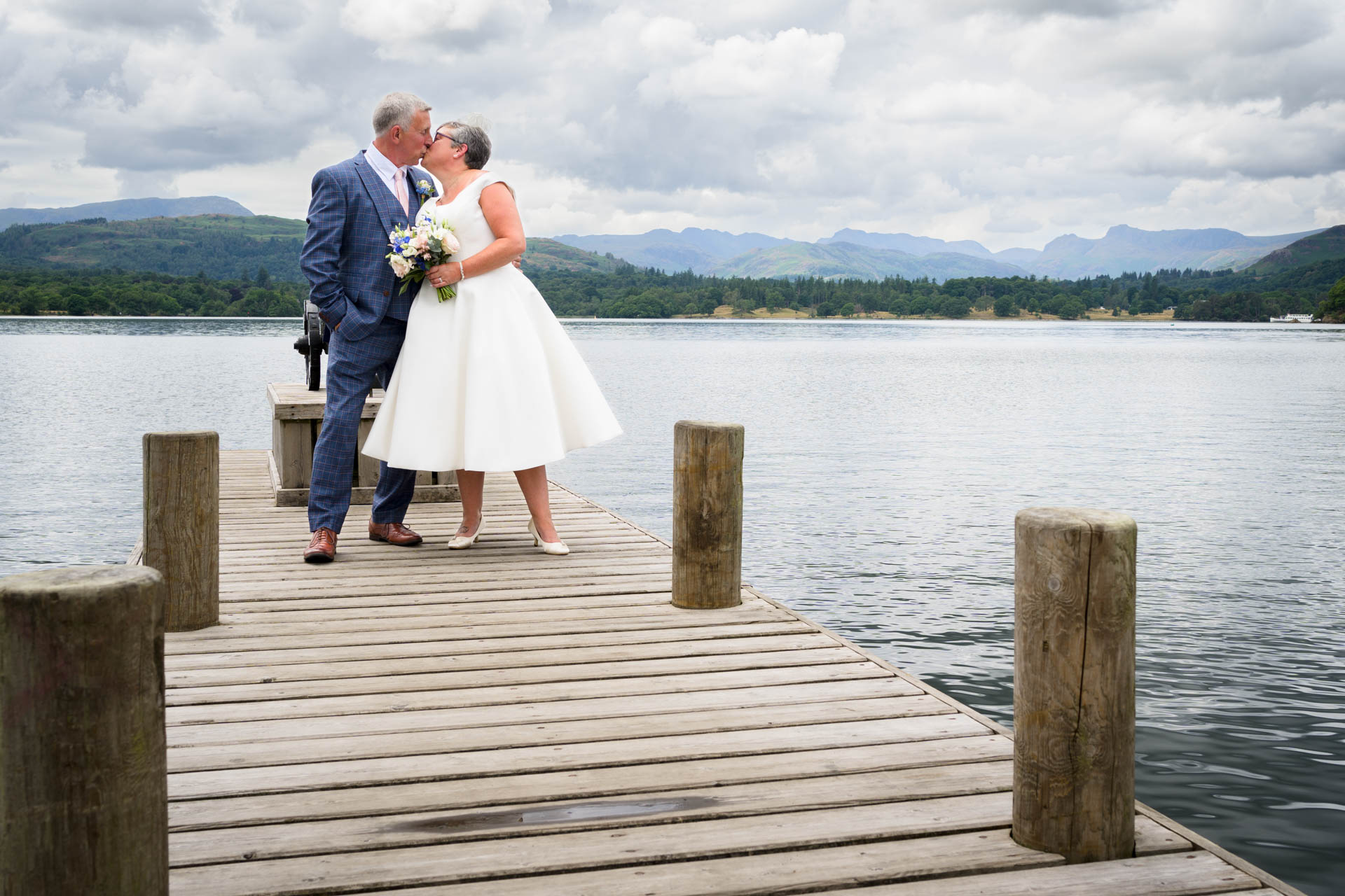 Wedding portrait of bride and groom kissing on jetty at Windermere with lake and mountains in background.