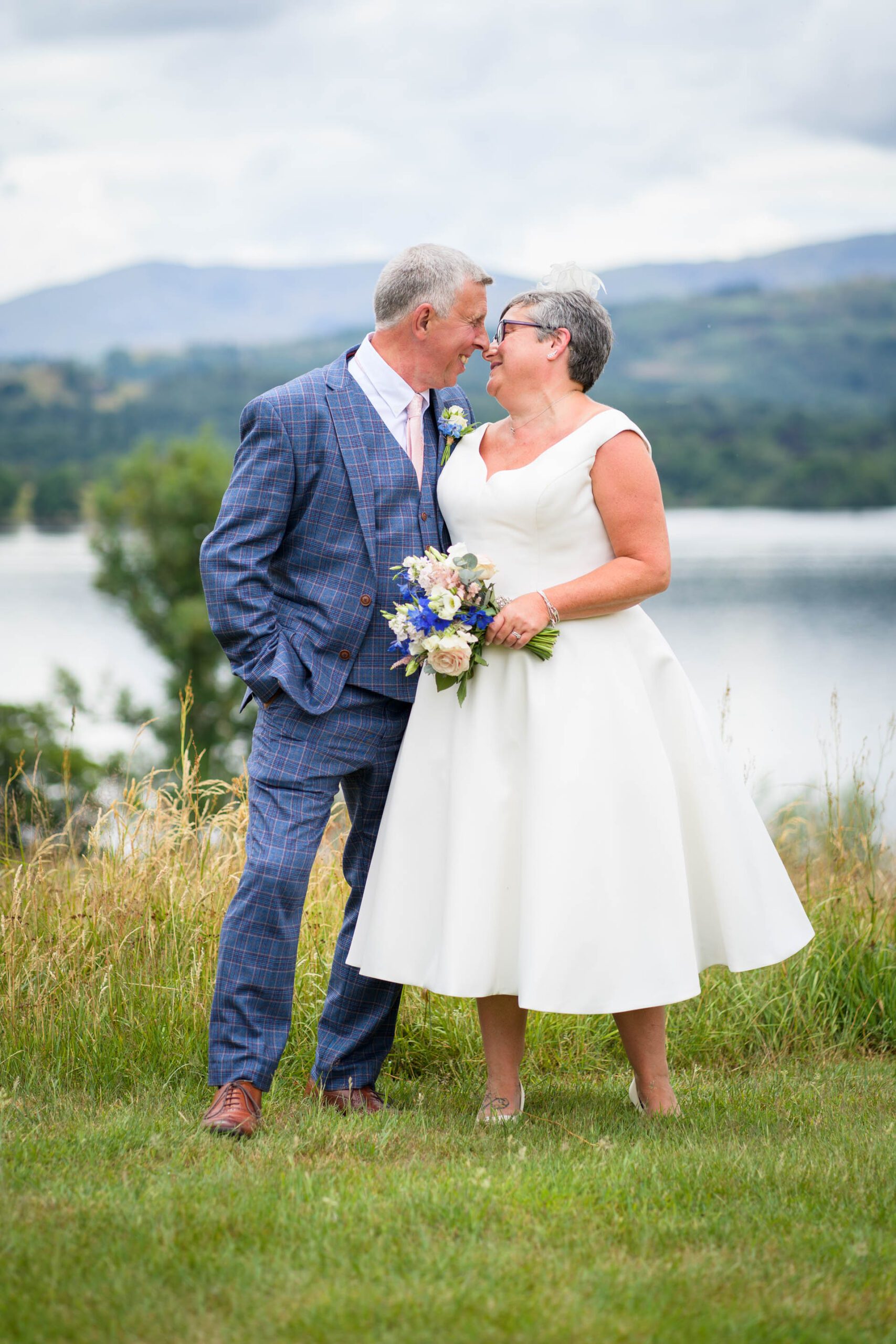 Wedding portrait of bride and groom on hill above Windermere with lake and mountains in background. Taken at their Low Wood Bay Resort wedding.