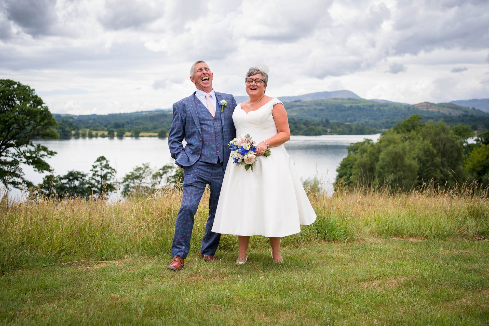 Wedding portrait of bride and groom on hill above Windermere with lake and mountains in background. 