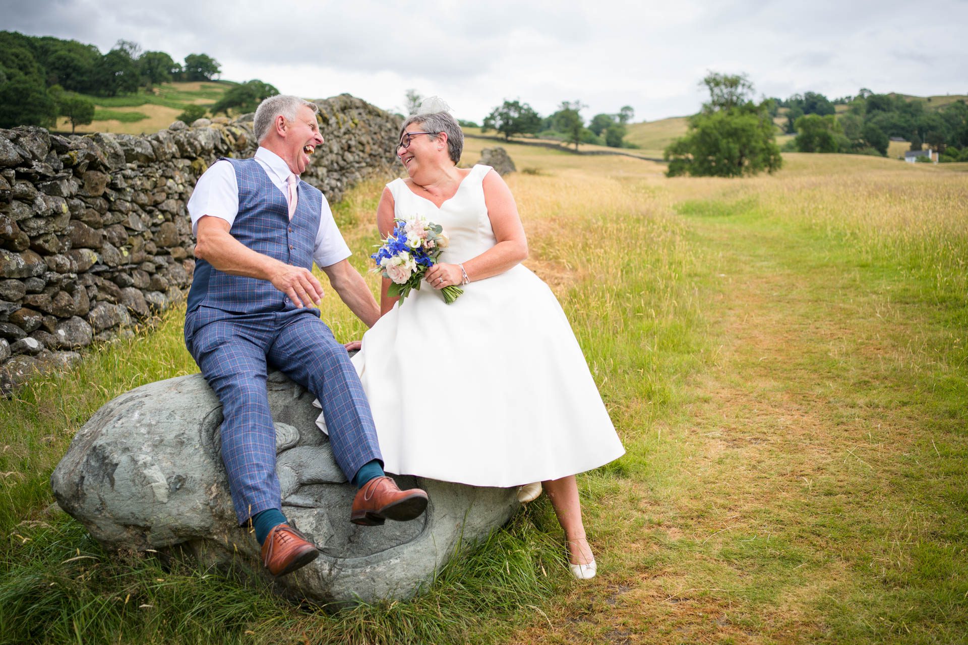 Bride and groom sitting on rock laughing. Dry stone wall, meadow and rolling hills in background. Taken at their Low Wood Bay Resort wedding.