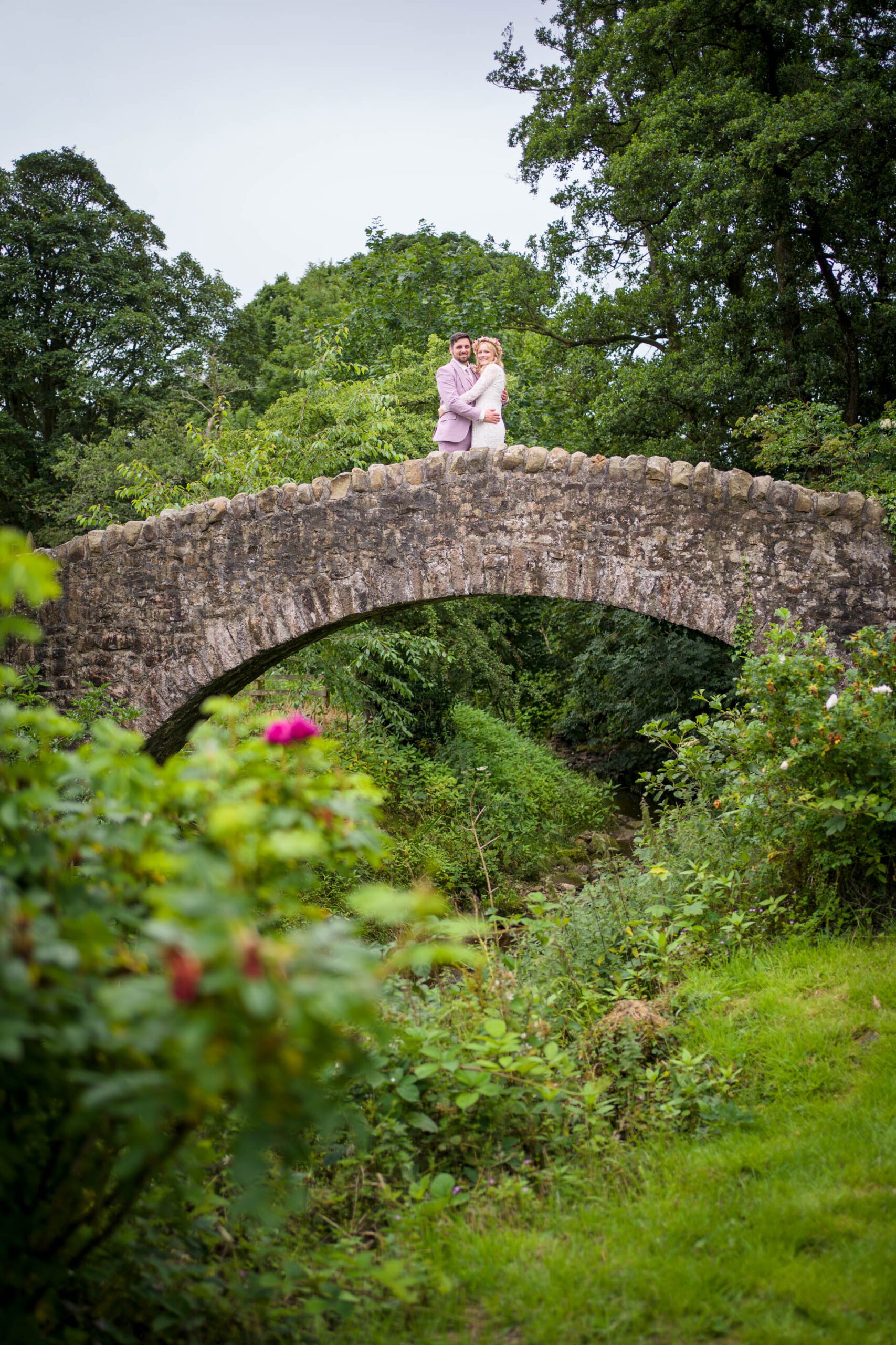 Wedding portrait at Ribble Valley wedding - bride and groom hugging while standing in centre of ancient arched stone bridge 